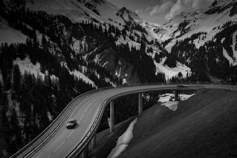 Dramatic view of cars on a winding highway through snow-covered Alpine mountains in black and white.
